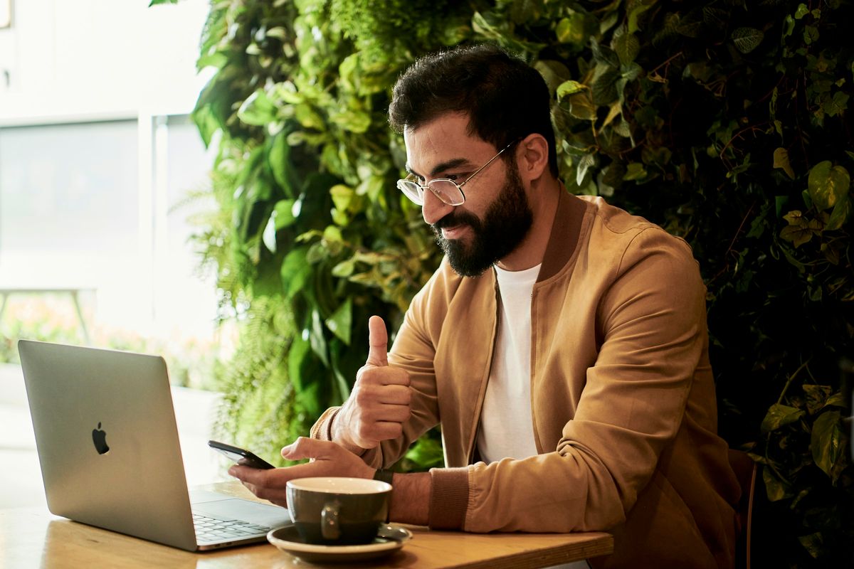 A smiling man giving a thumbs up at his laptop in a warm café